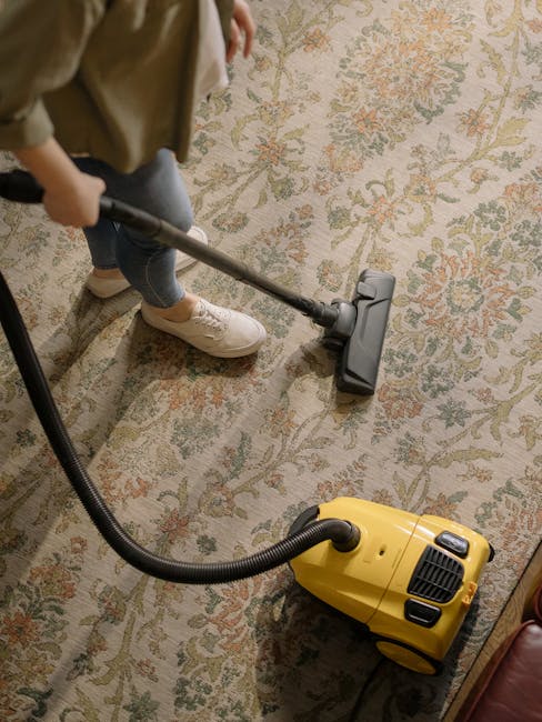 A woman vacuuming a patterned area rug in a living room with wooden flooring and a leather sofa. She is using a yellow canister vacuum cleaner, focusing on surface cleaning to remove dust and debris f