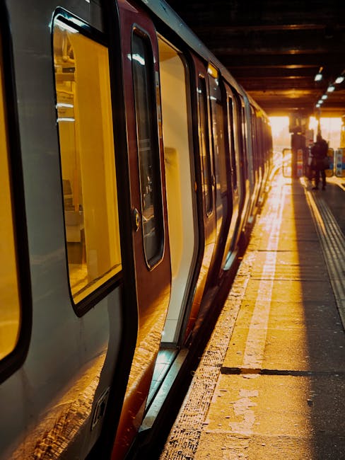 A fast-moving train inside a railway station with a glass and steel roof structure overhead. The train features a red, white, and blue colour scheme, appearing blurred due to motion. The station wall 
